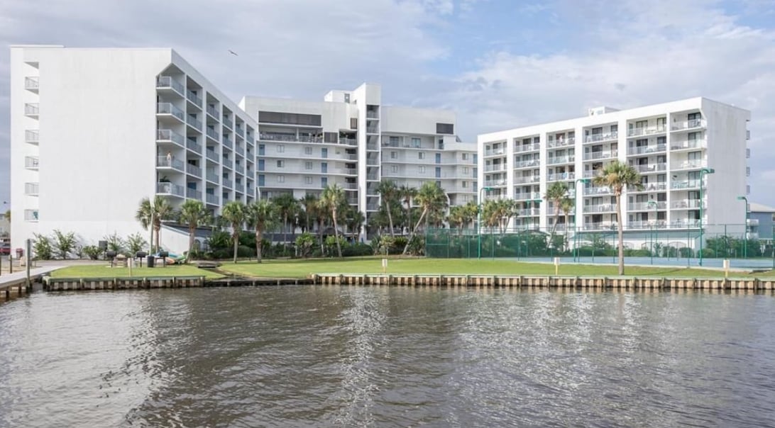 A modern condo complex is visible against a cloudy sky, featuring multiple levels and balconies. Manicured lawns with palm trees surround the building. Water reflects the structure, suggesting proximity to a lagoon or bay. Tennis courts can be seen in the background.
