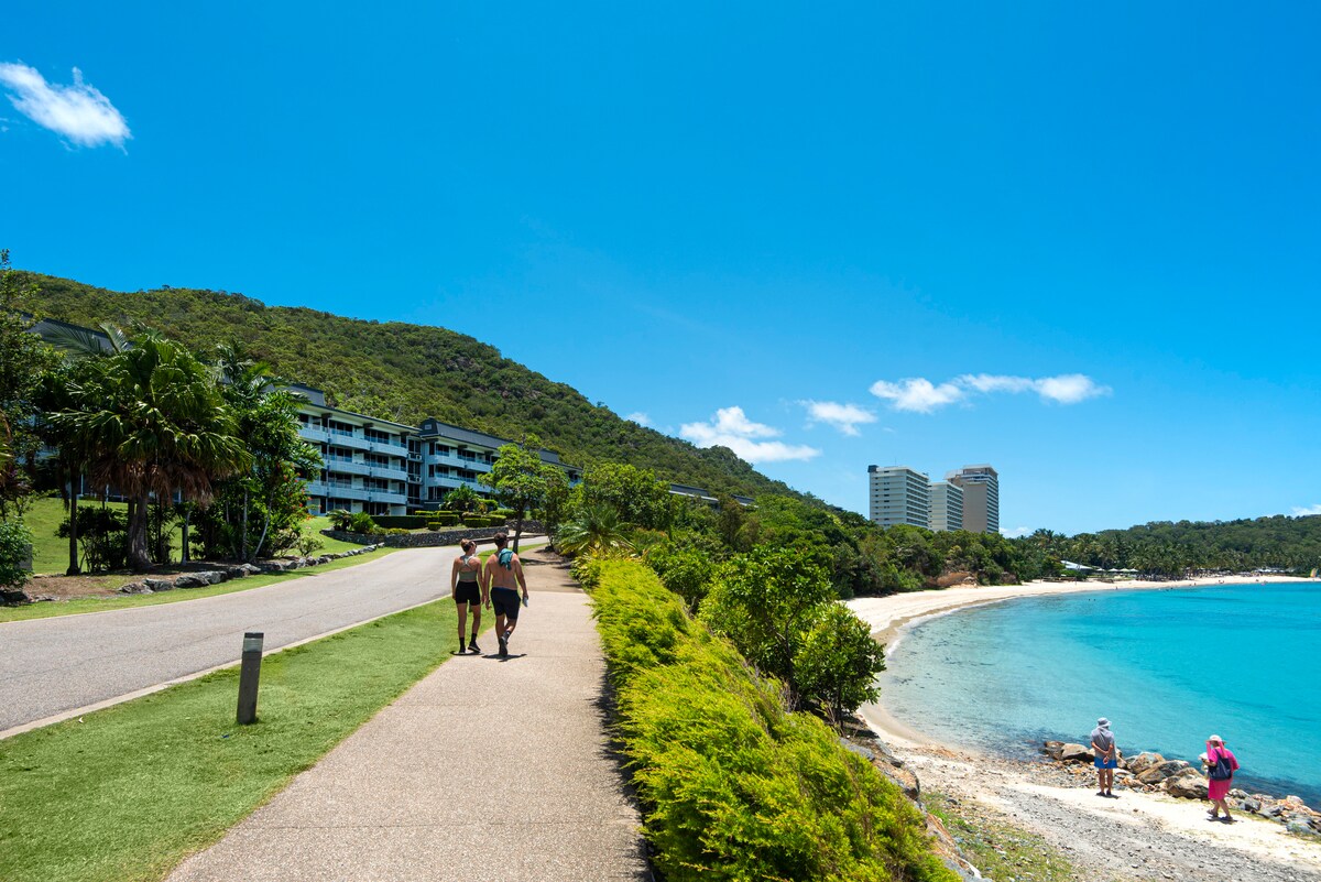A scenic pathway runs alongside a serene beach, with lush greenery and gentle hills in the background. Pedestrians stroll leisurely, enjoying the coastal view. The clear blue sky complements the tranquil ocean, while resort buildings are visible in the distance, creating a harmonious blend of nature and luxury.