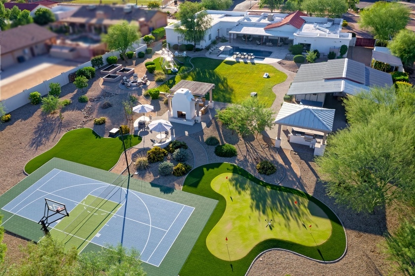 An aerial view reveals a spacious backyard oasis featuring a basketball court, putting green, and landscaped areas with seating. Various shaded spots are visible, including a gazebo and outdoor dining area, all surrounded by greenery and designed for relaxation and recreation.