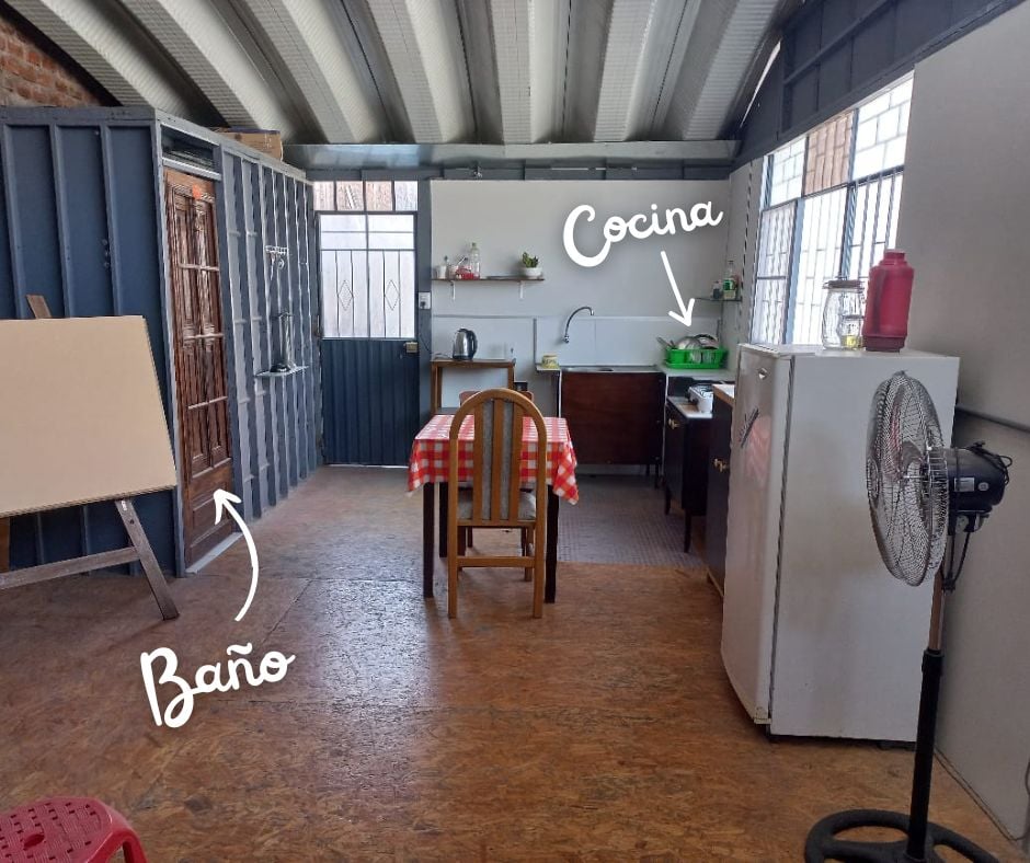A shared space features a dining area with a checkered tablecloth and a small kitchen area equipped with essential appliances. A refrigerator stands adjacent to the cooking space, while the bathroom is indicated with a directional label. Natural light fills the room from windows.