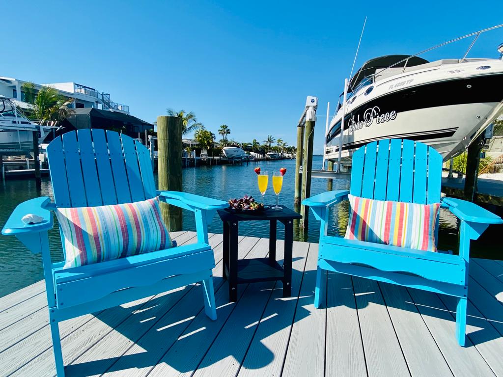 Two vibrant blue Adirondack chairs with striped cushions are positioned on a wooden dock overlooking a canal. A small black table holds two drinks, providing a relaxing setting. A docked boat is visible in the background, with clear blue skies reflecting on the water.