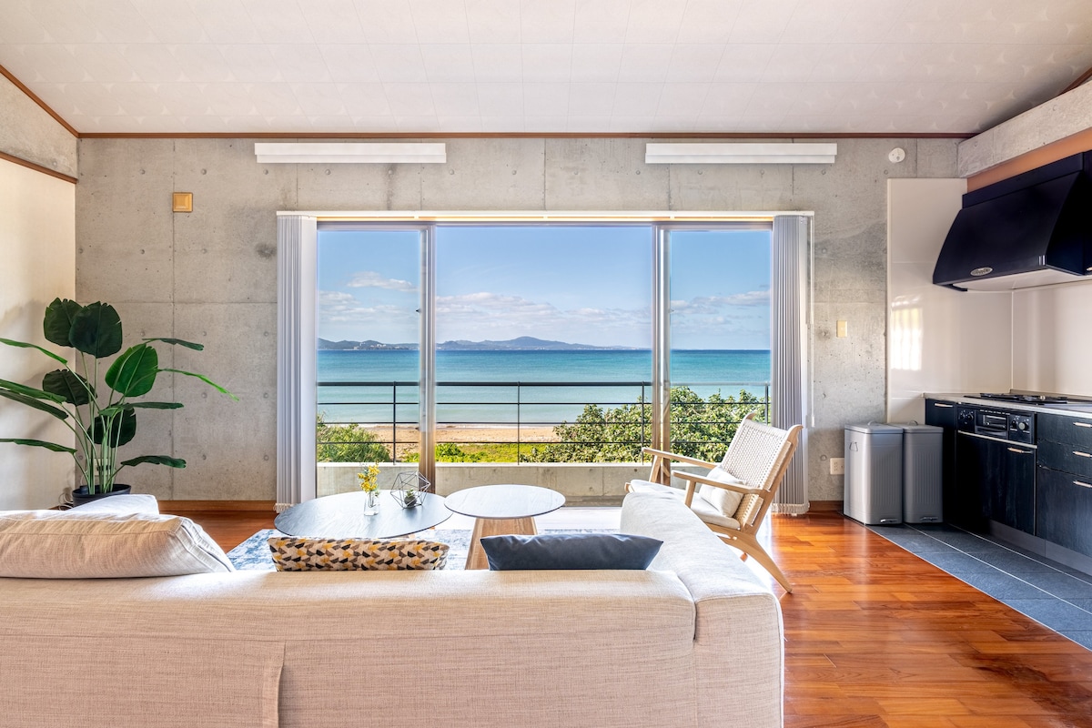 The spacious living area features large windows offering a clear ocean view. Natural light floods the room, highlighting the contrast of the wooden floor against the concrete walls. A white sofa and a wooden chair provide comfortable seating, with a small table displaying a flower vase.