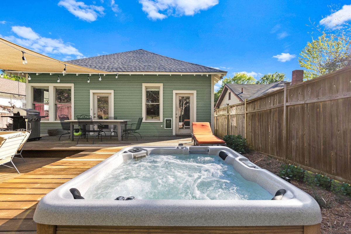 A relaxing outdoor space features a hot tub with bubbling water, surrounded by a wooden deck. Sun loungers and a dining area with a table are visible, set against a backdrop of a charming green house. A shaded area is provided by a large canopy.