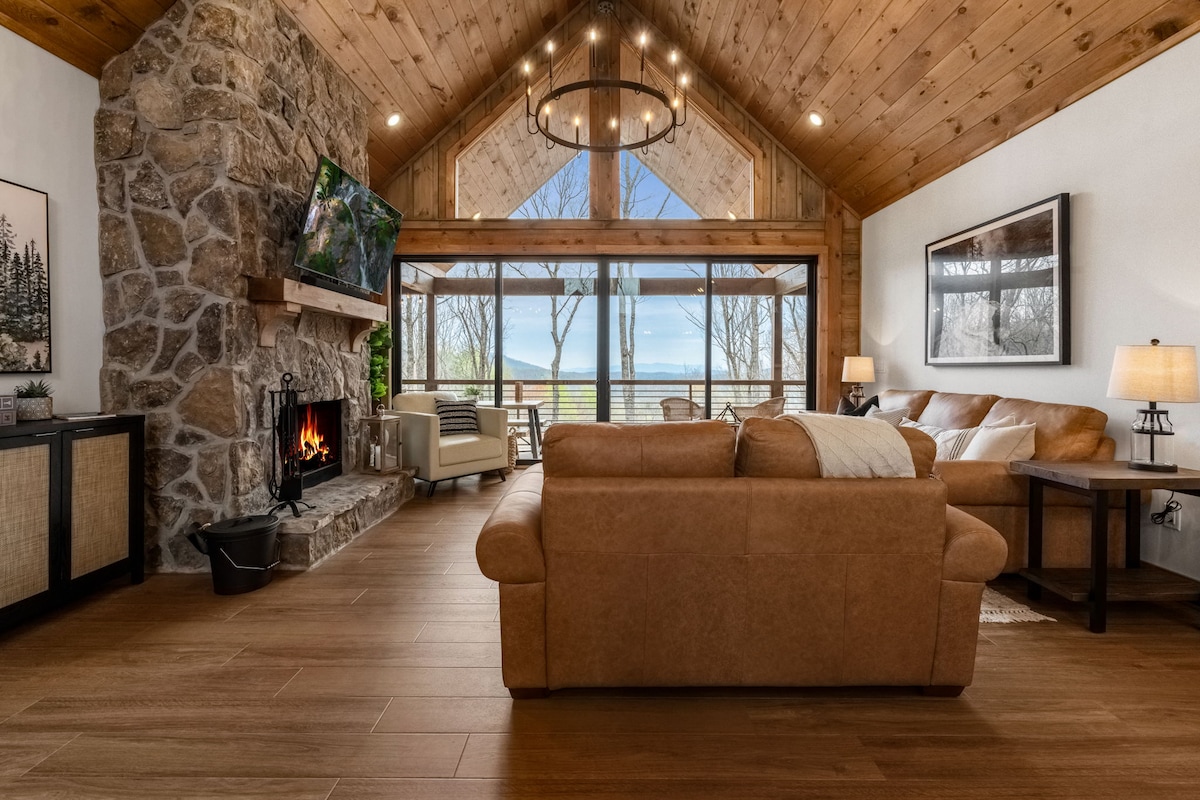 A spacious living area features natural wood accents and large windows that frame panoramic views of the mountains. A stone fireplace sits beside a flat-screen TV, and comfortable leather couches provide seating for relaxation. Soft lighting adds warmth to the inviting atmosphere.