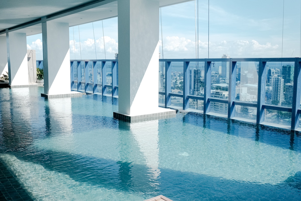 A tranquil swimming pool is shown, featuring clear water reflecting the sky and cityscape. Large glass windows provide a panoramic view. The area is well-lit, creating a serene atmosphere. The pool is complemented by modern architectural elements and open spaces.