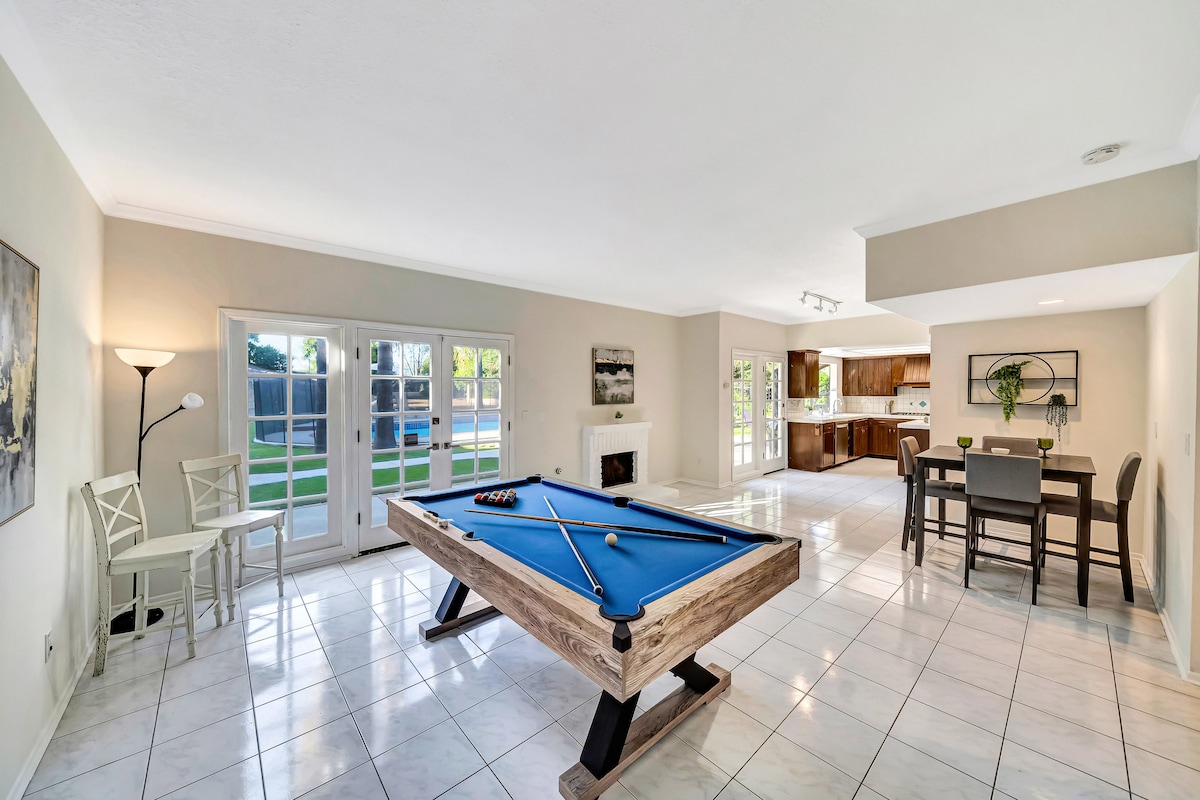The airy living space is highlighted by a billiard table and a cozy seating area. Large glass doors provide ample natural light and views of the outdoor space. A second fireplace is positioned nearby, with an elegant dining area visible in the background.