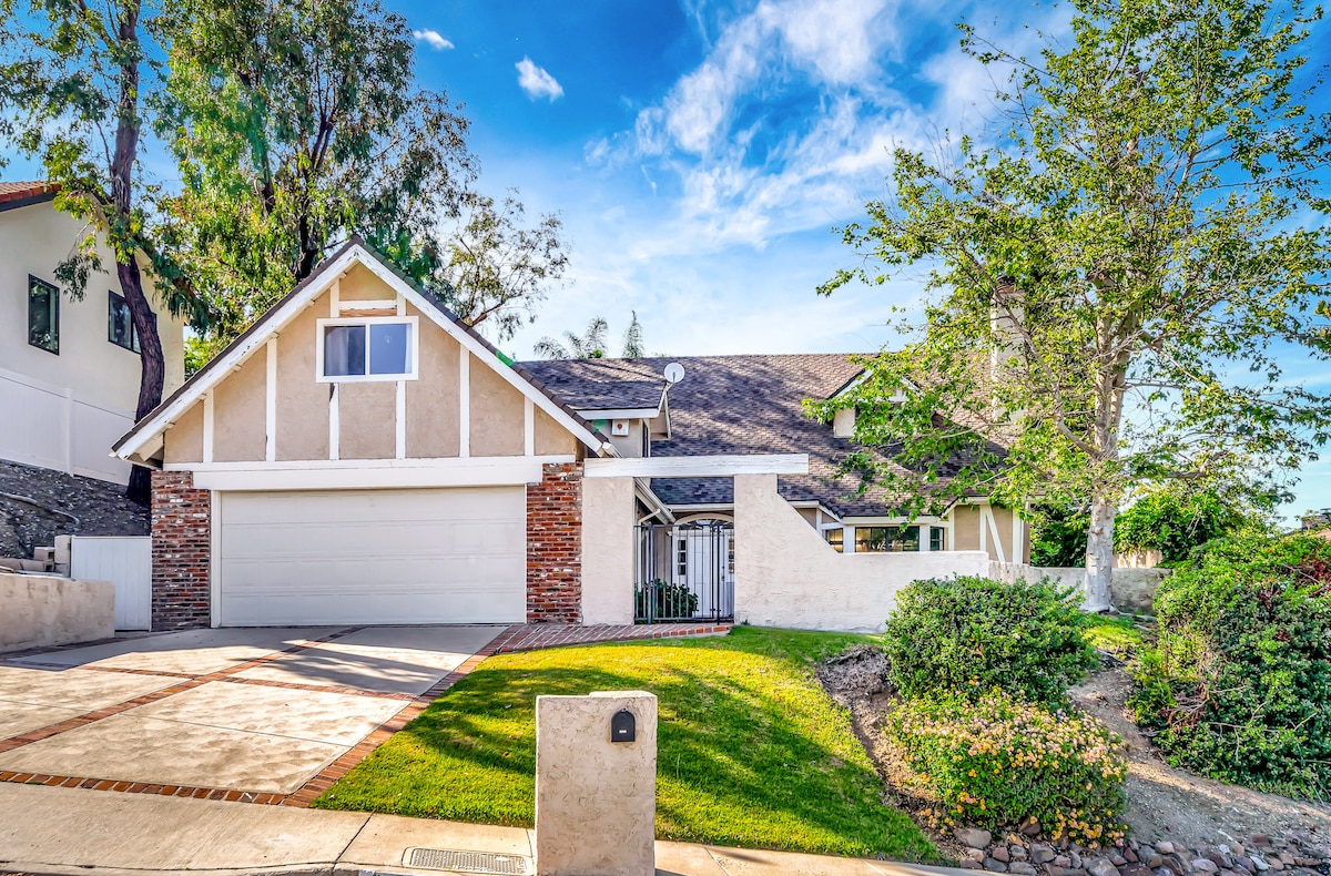 The exterior of the house showcases a two-story structure with a combination of textured light-colored walls and a sloped roof. A well-maintained lawn surrounds the property, complemented by a driveway and a stone path leading to the entrance. Lush trees and greenery are visible.