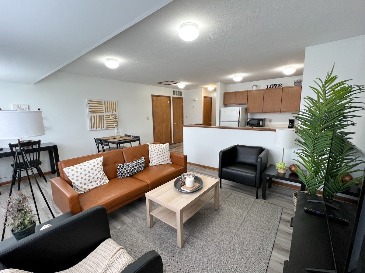 A comfortable living area features a brown leather sofa with decorative cushions and two black armchairs. A light wooden coffee table is set at the center, with a small plant. In the background, a dining area and kitchen are visible, providing a functional open layout.
