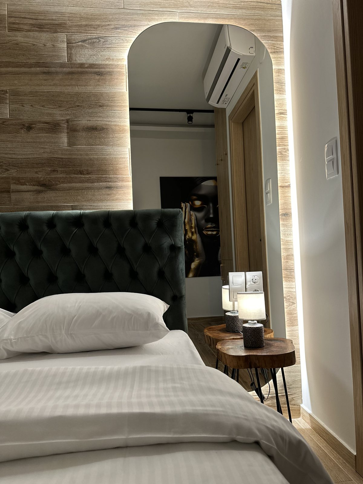 A stylish bedroom is featured with a tufted headboard and a well-made bed. Soft lighting is reflected in a decorative mirror, enhancing the contemporary decor. Two wooden side tables hold candles, adding a touch of elegance to the space.