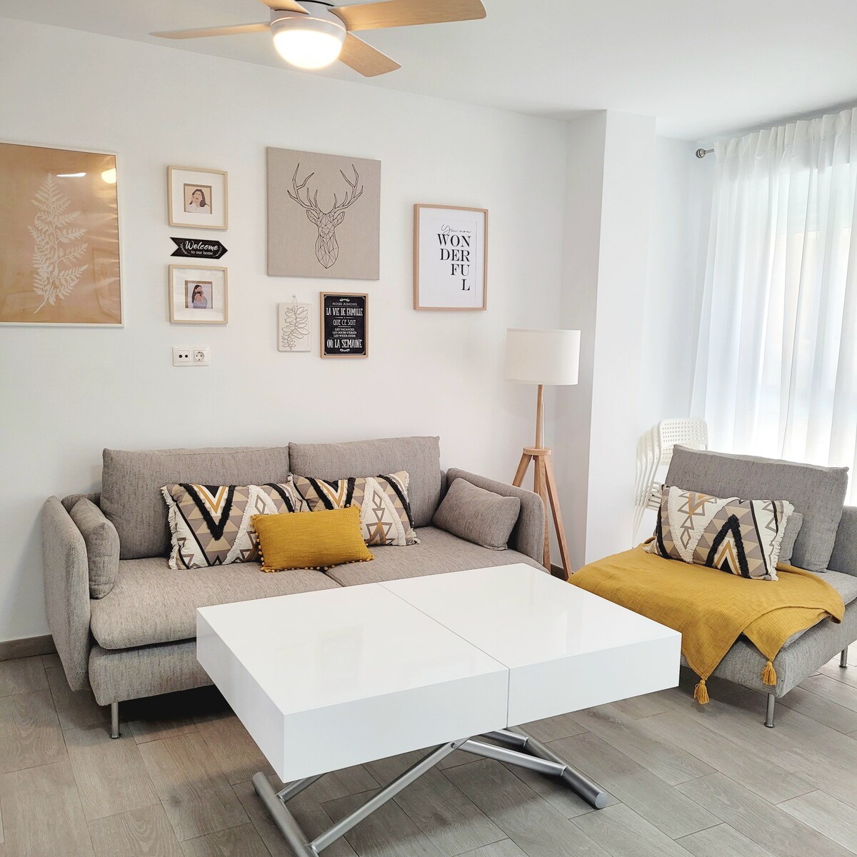 A modern living area is featured, highlighting a gray sectional sofa adorned with patterned cushions and a yellow throw pillow. A light wood fan is visible above. The decor includes framed art on the wall, and a white coffee table with a unique design is centrally placed.