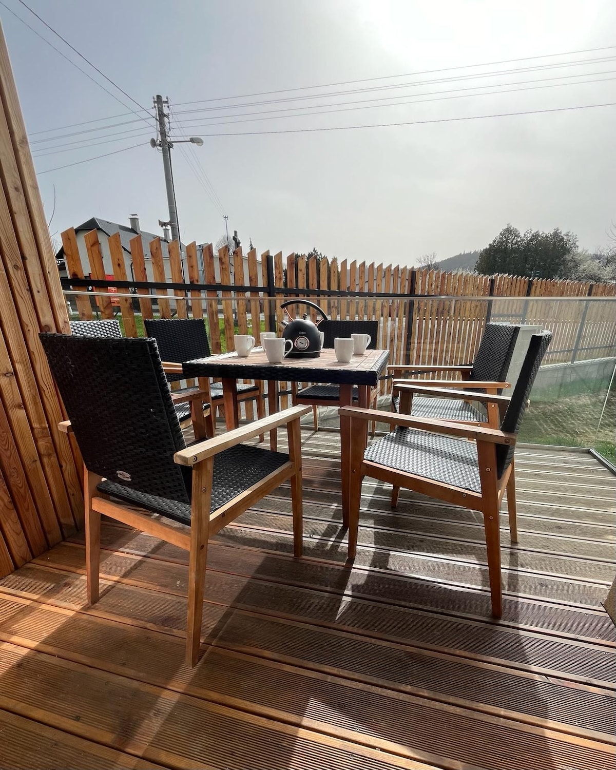 A small outdoor seating area is presented, featuring a wooden table surrounded by four wicker chairs. A kettle and a set of cups are placed on the table. The space is enclosed by a wooden fence, providing privacy.