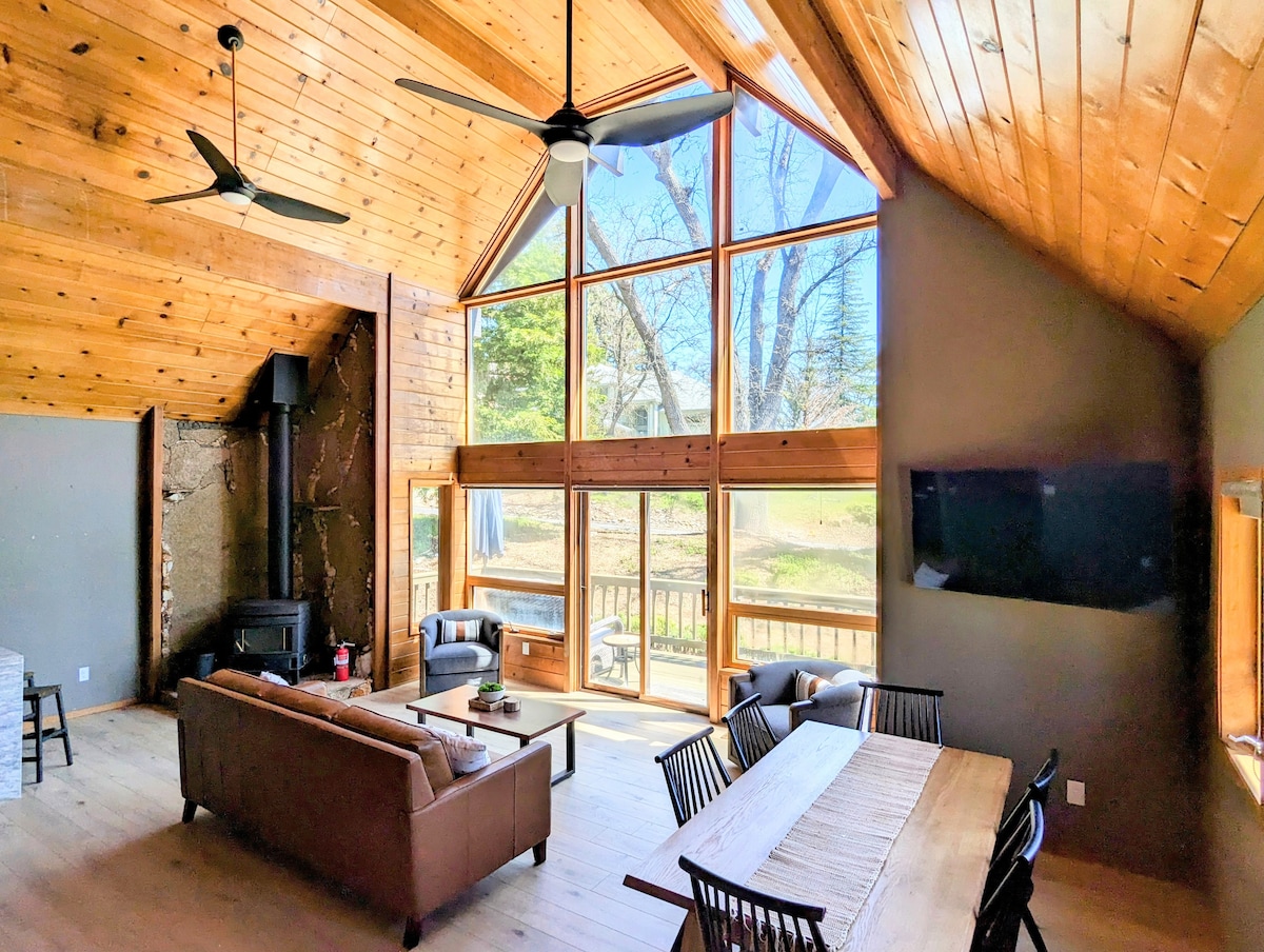 The spacious living area features large floor-to-ceiling windows, allowing natural light to fill the space. A plush leather sofa and wooden dining table for six are positioned beneath a vaulted wooden ceiling, creating a cozy yet open atmosphere. A flat-screen TV is mounted on the wall.