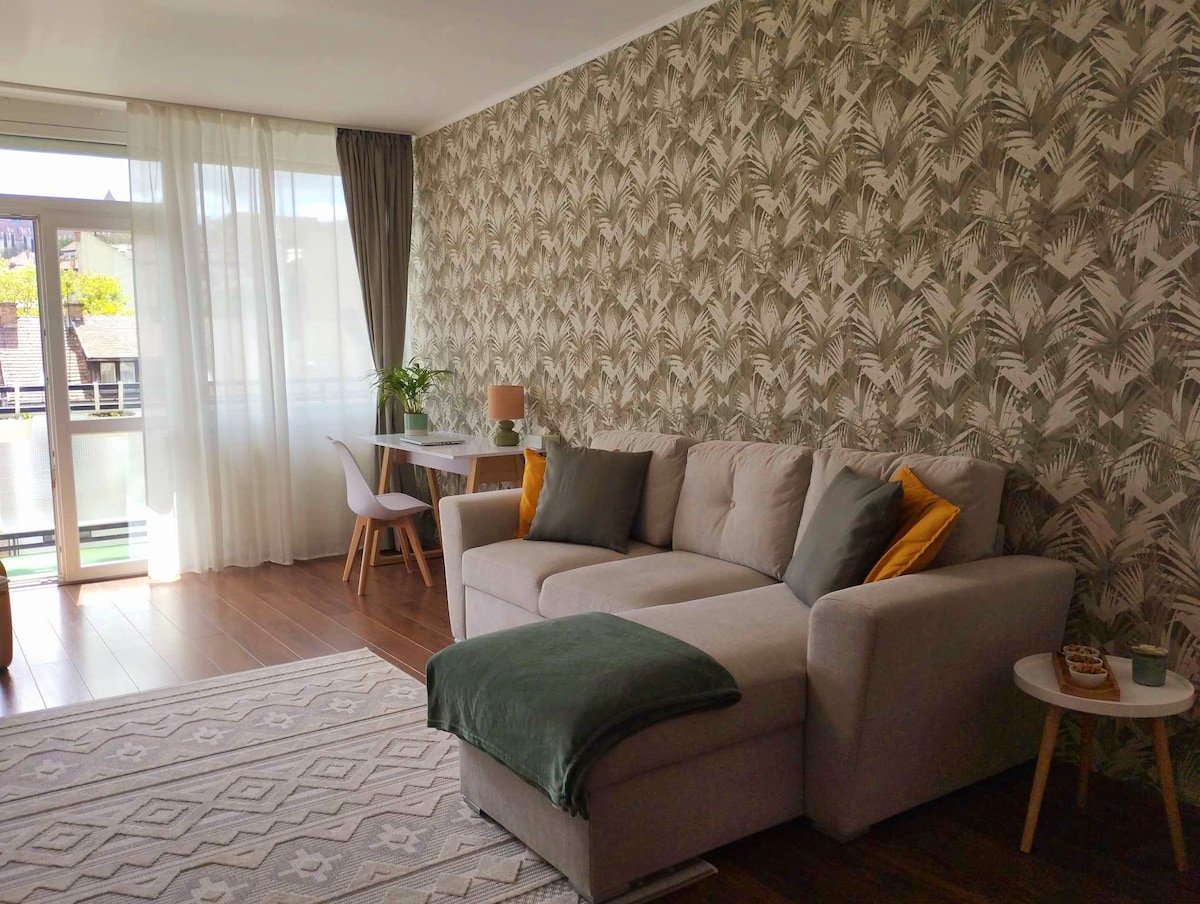 A comfortable living area is highlighted by a light-colored sofa with accent pillows. A small table and chair provide a cozy dining space, while large windows allow natural light to fill the room. Decorative wallpaper with a tropical motif adds a vibrant touch.