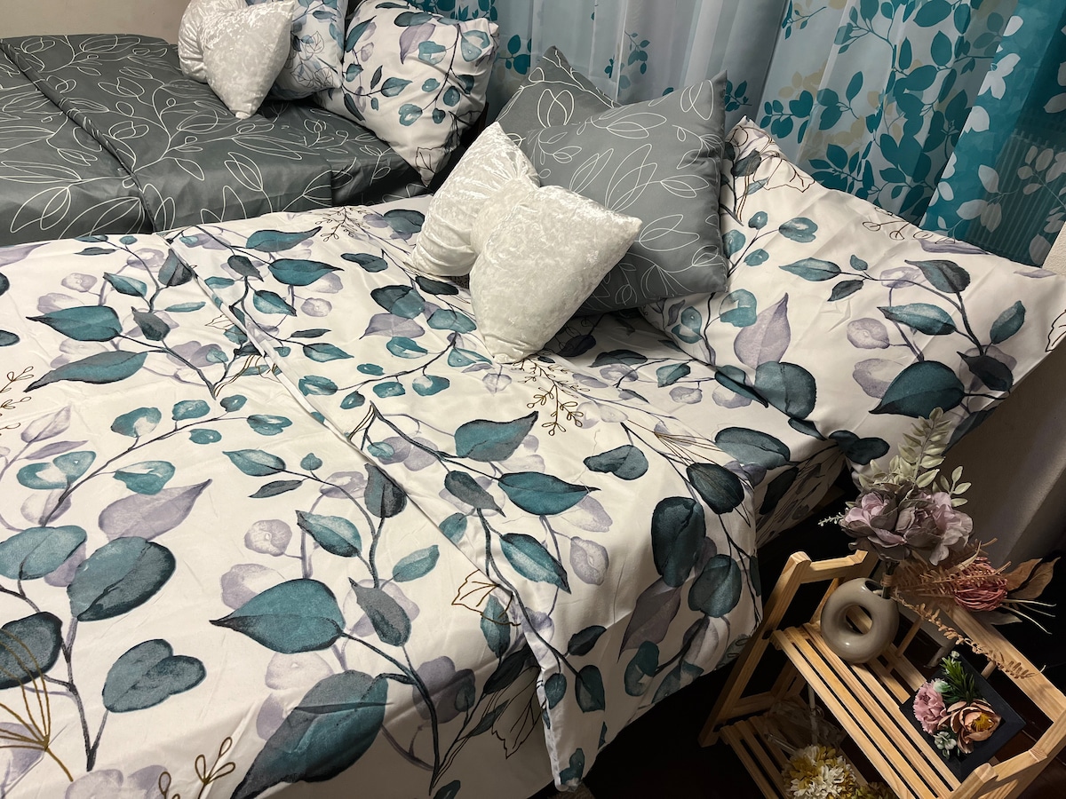 A room featuring two separate beds adorned with floral-patterned bed linens in shades of teal and gray. Cushions in varying designs, including white plush and patterned, are arranged on and around the beds. A wooden shelf holds decorative items, complementing the serene atmosphere.