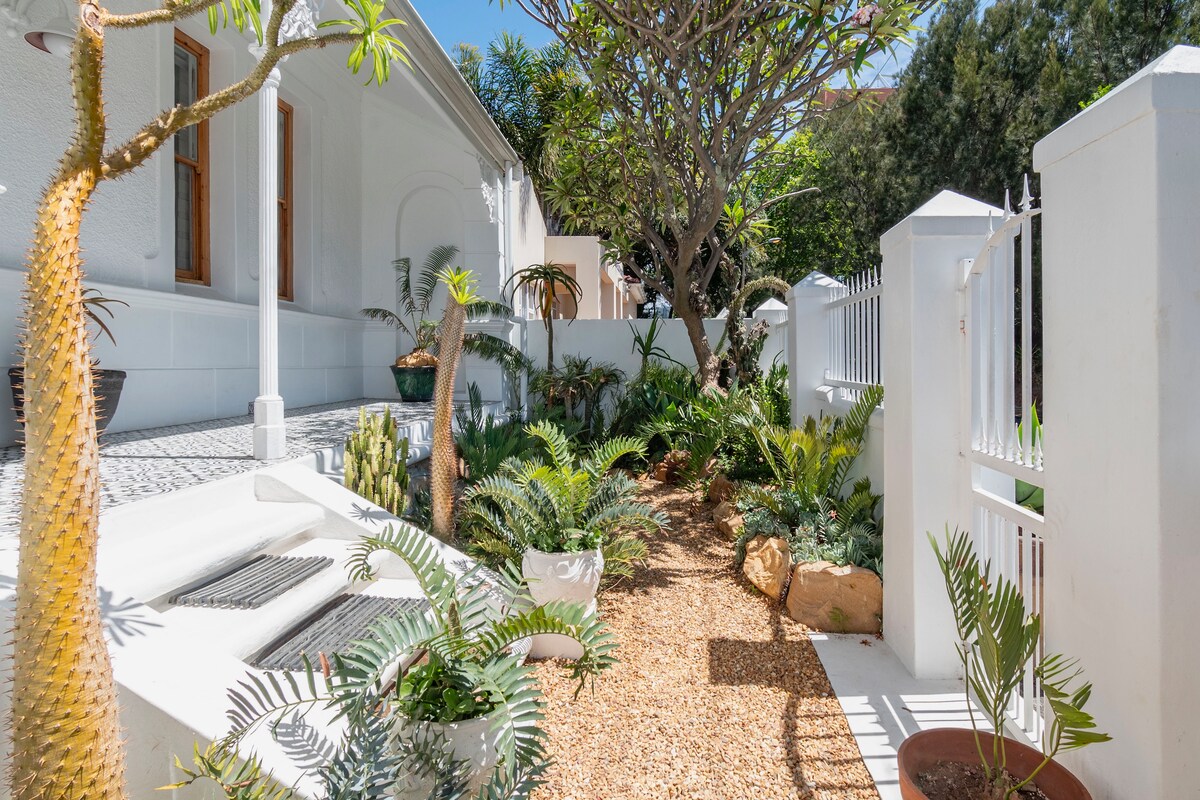 A garden pathway leads through a lush arrangement of greenery, featuring various plants in decorative pots. Natural textures are highlighted by pebbled ground and soft sunlight filtering through the trees, enhancing the serene ambiance of this outdoor space.
