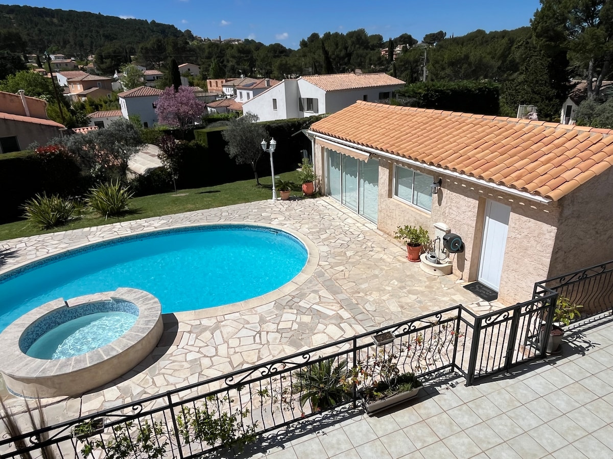 A private pool area features a circular pool and a spa section, surrounded by a stone patio. The outdoor space is framed by neatly trimmed hedges and greenery, with a spacious deck providing a view of the tranquil landscape and nearby houses.