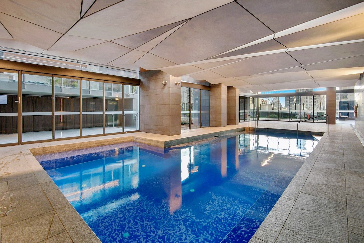 An indoor swimming pool is featured, surrounded by sleek stone walls and large glass windows that provide a view of the outdoors. The water is crystal clear, reflecting the modern architectural design overhead. Soft lighting creates a calm atmosphere.