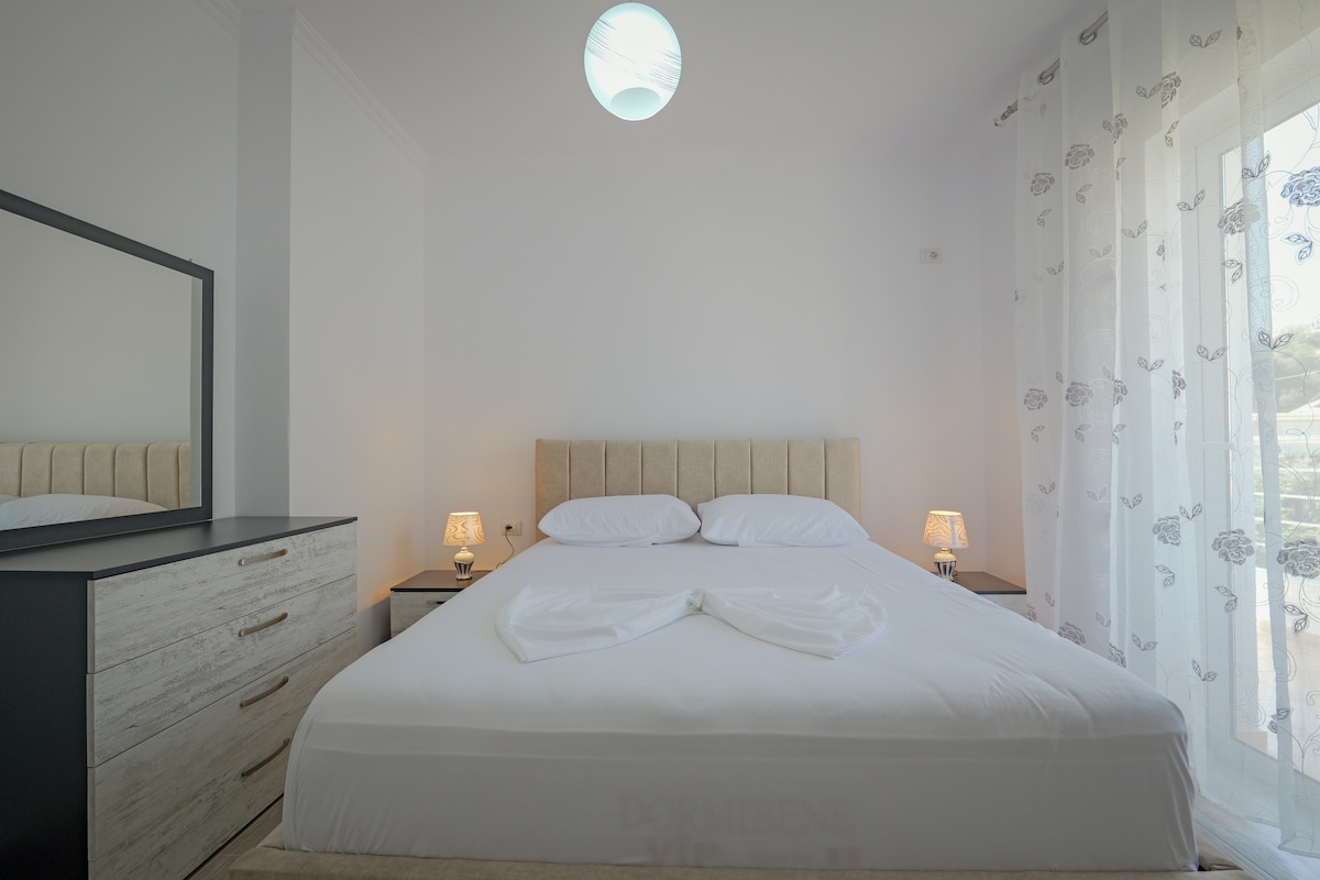 A spacious bedroom is highlighted by a plush bed, neatly made with fresh linens. Soft lighting from bedside lamps and a ceiling fixture enhances the atmosphere. A dresser and a light window curtain complement the decor, while a portion of the balcony is visible through glass doors.