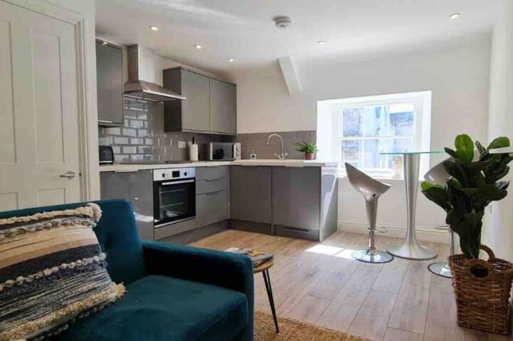 A contemporary kitchen features sleek cabinetry and modern appliances. A small round glass table is accompanied by two stylish stools. A cozy blue sofa is positioned nearby, accentuated by textured cushions, with natural light pouring in through the window.