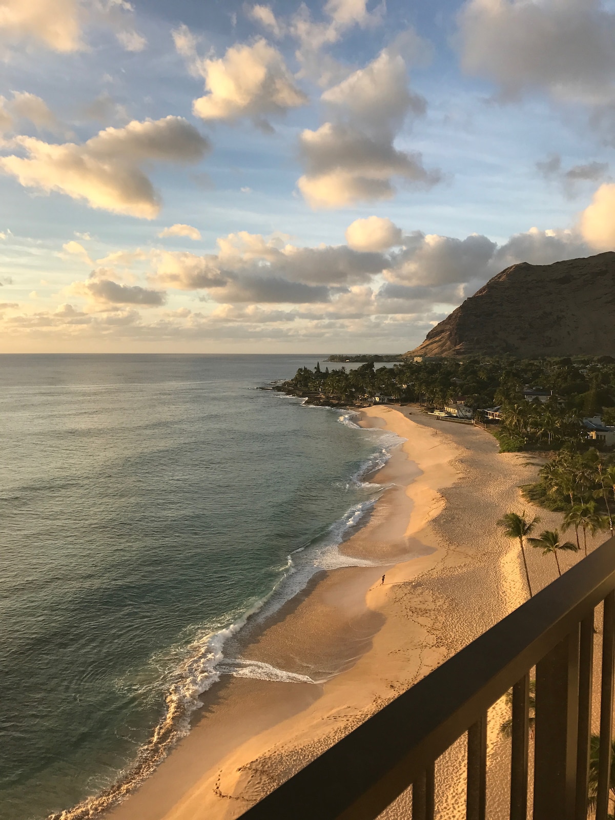 A tranquil beach scene captures the gentle curves of a sandy shoreline meeting the calm ocean. Soft waves caress the shore under a sky painted with clouds at sunset. Nearby, a lush mountain rises, creating a serene backdrop to the sparsely populated beach.