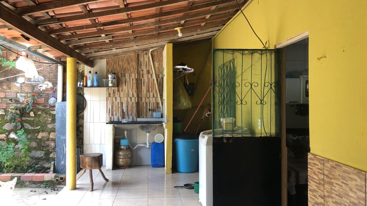 An open living space features yellow walls and a tiled floor. A wooden table is positioned in the foreground, and a blue water container is visible. Storage racks hold various items, and a small window with a decorative iron grille allows natural light into the area.