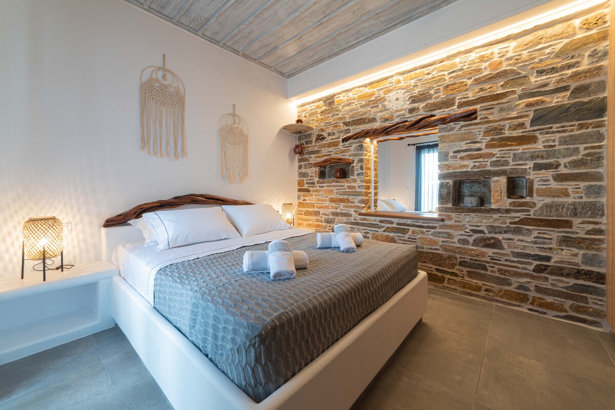 A spacious bedroom features a double bed dressed in neutral tones, complemented by decorative wall hangings. Soft lighting accentuates the textured stone wall and wooden mirror. Fresh towels are neatly arranged on the bed, providing a welcoming touch.