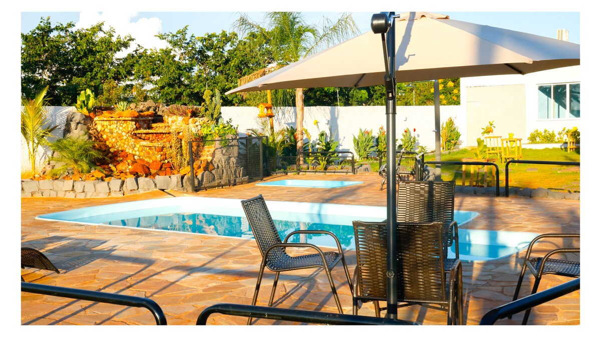 Two swimming pools are set within the spacious outdoor area, surrounded by lush greenery and decorative landscaping. A shaded seating area with several black chairs and a large umbrella offers comfort. A serene waterfall feature complements the relaxing ambiance of the environment.