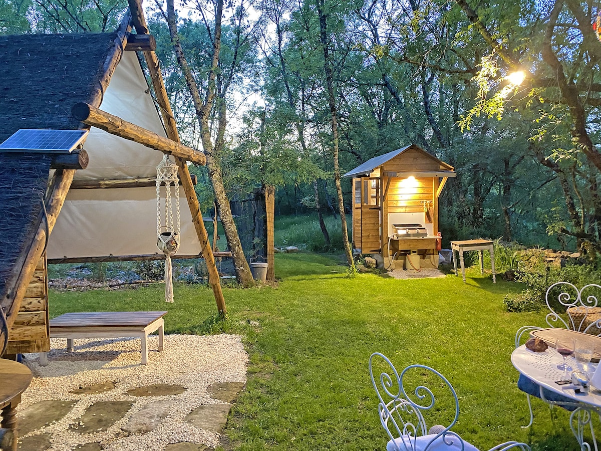 An outdoor area is shown, featuring a unique cabin beside a small wooden kitchen shed. A swing hangs nearby, and a seating arrangement is placed on a stone-paved section, surrounded by lush greenery. Soft lighting from the kitchen adds a warm glow to the scene.