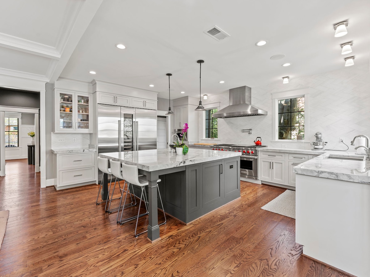 The spacious kitchen features a large island with seating and premium appliances, including a stainless steel refrigerator and an elegant vent hood. White cabinetry and marble countertops complement the warm wood flooring, while natural light floods in through a window.