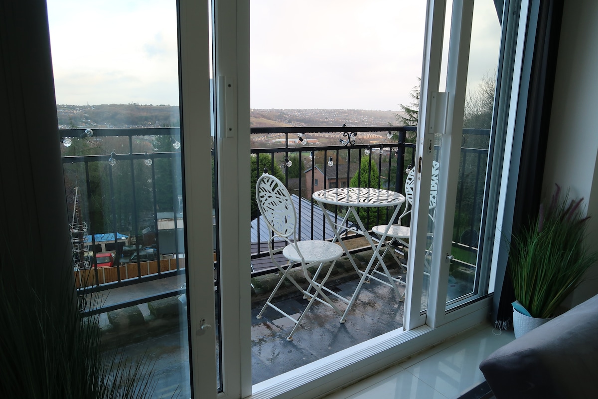 A balcony area features a small round table with two chairs, overlooking a scenic view of the surrounding landscape. Glass doors provide access, with greenery visible on either side, creating a bright and airy atmosphere.