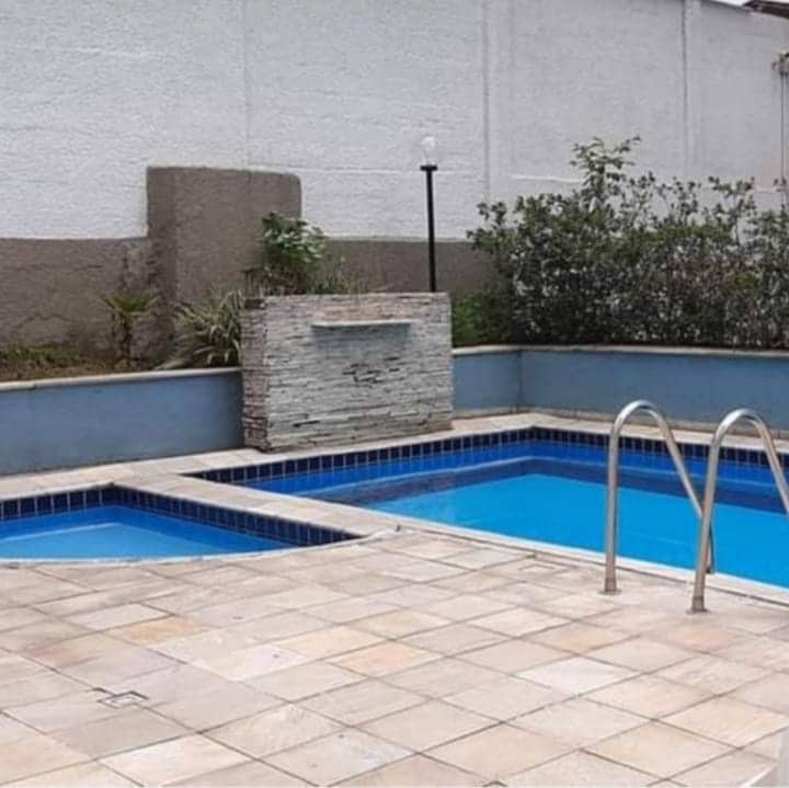 A pool area is featured, consisting of two rectangular pools, one designed for adults and the other for children. Surrounding the pools are light-colored tiles and greenery, with a stone water feature creating a calming element. A lamp post stands nearby, enhancing the ambiance.