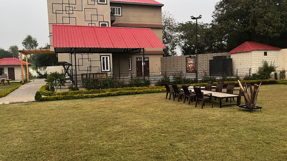 A spacious outdoor area features a neatly manicured lawn, complemented by a long dining table surrounded by several chairs. A two-story building with a red roof is visible in the background, along with decorative planters and trees that enhance the natural setting.
