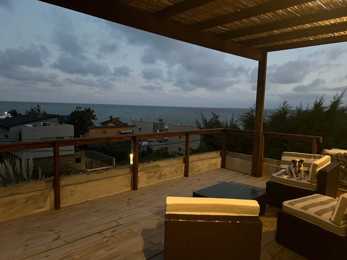 An expansive terrace is captured at twilight, featuring comfortable seating areas overlooking the ocean. The gentle waves create a serene backdrop, while the wooden deck is illuminated softly, enhancing the inviting space for relaxation.