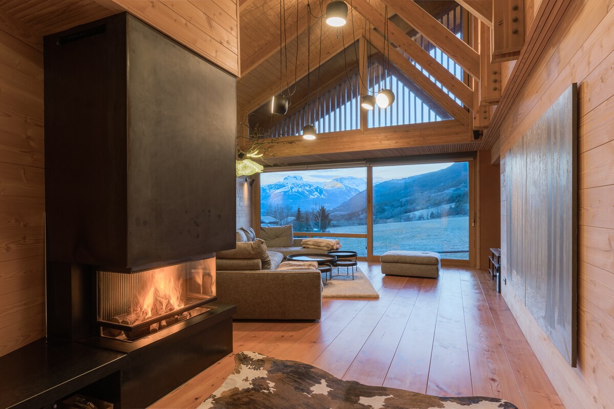A spacious living area features large windows showcasing mountain views. A modern fireplace is positioned centrally, with a cozy seating arrangement surrounding it. The warm wooden walls and flooring contribute to a calming ambiance, accentuated by stylish overhead lighting.