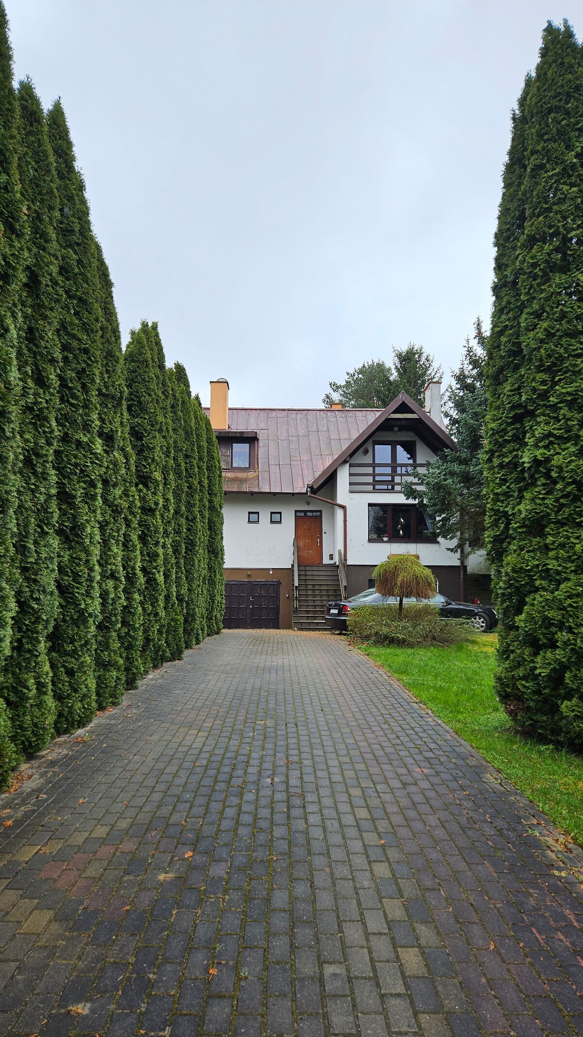 A tree-lined driveway offers a view of a two-story house with a sloped roof, surrounded by tall evergreens. The path leads toward a welcoming entrance, featuring a wooden door framed by windows. A garage is situated on the left side of the house.