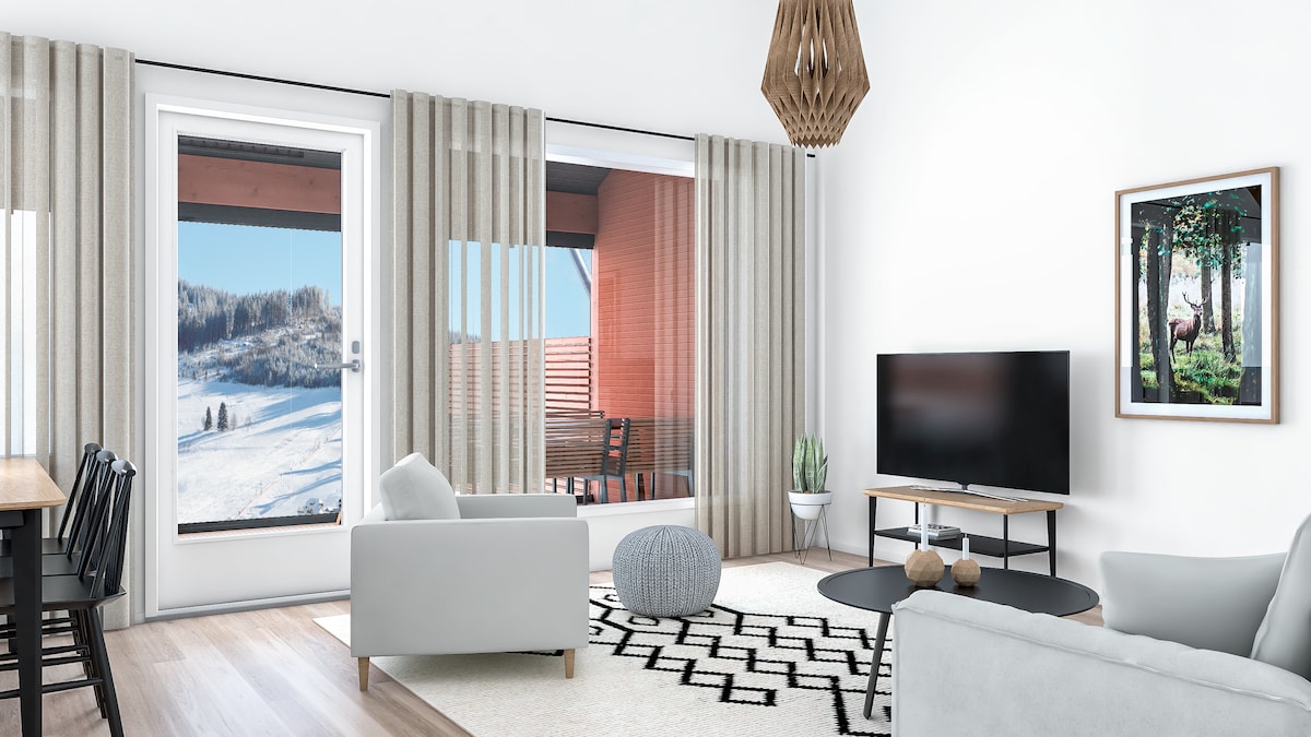 A bright living area features large glass doors leading to a snowy outdoor view. Light-colored curtains frame the windows. A comfortable sofa and armchair sit on a textured area rug. A modern television is positioned on a console against the wall, accompanied by a decorative artwork.