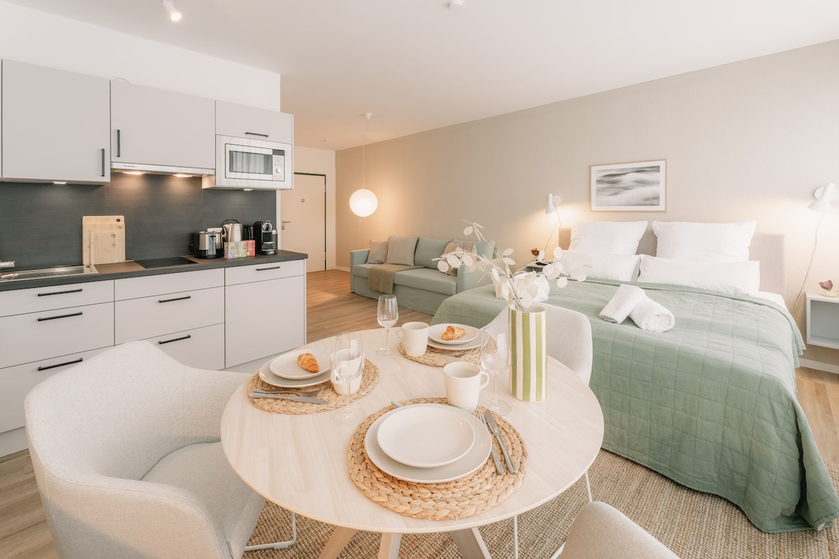 A modern studio apartment features a well-equipped kitchen and a dining area with a circular table set for two. A comfortable seating area is visible, alongside a spacious bed with crisp linens. Soft lighting enhances the inviting atmosphere, and neutral tones create a cohesive look.