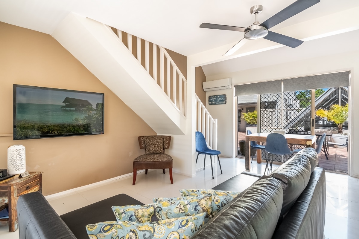A modern living area features a large flat-screen TV mounted on the wall, along with a comfortable leather sofa adorned with decorative cushions. Natural light streams through nearby glass doors that open to an outdoor dining space, complemented by a ceiling fan.