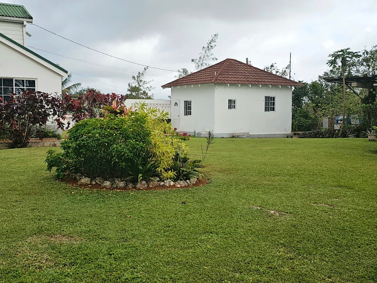 A well-kept garden area is shown, featuring manicured green grass and a circular flower bed with a variety of plants. The background reveals a white, single-story building with a red-tiled roof, surrounded by trees and shrubbery under a partly cloudy sky.