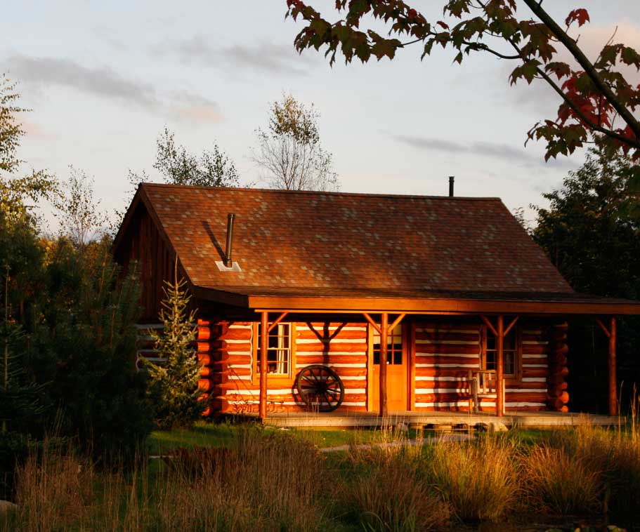 A rustic cabin is visible with a log exterior and a sloped roof. The entrance features a covered porch supported by wooden beams, with a wagon wheel displayed as decor. Surrounding greenery and a natural pond can be seen in the foreground, bathed in warm evening light.