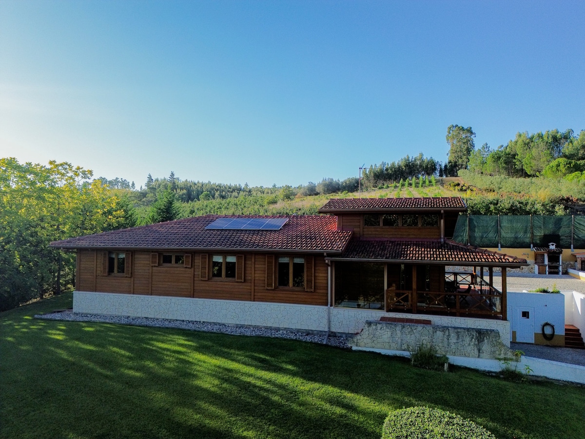 The exterior of a wooden house is presented, featuring a red-tiled roof and multiple windows under a clear blue sky. A well-maintained grassy area surrounds the property, while trees and gentle hills provide a serene backdrop.