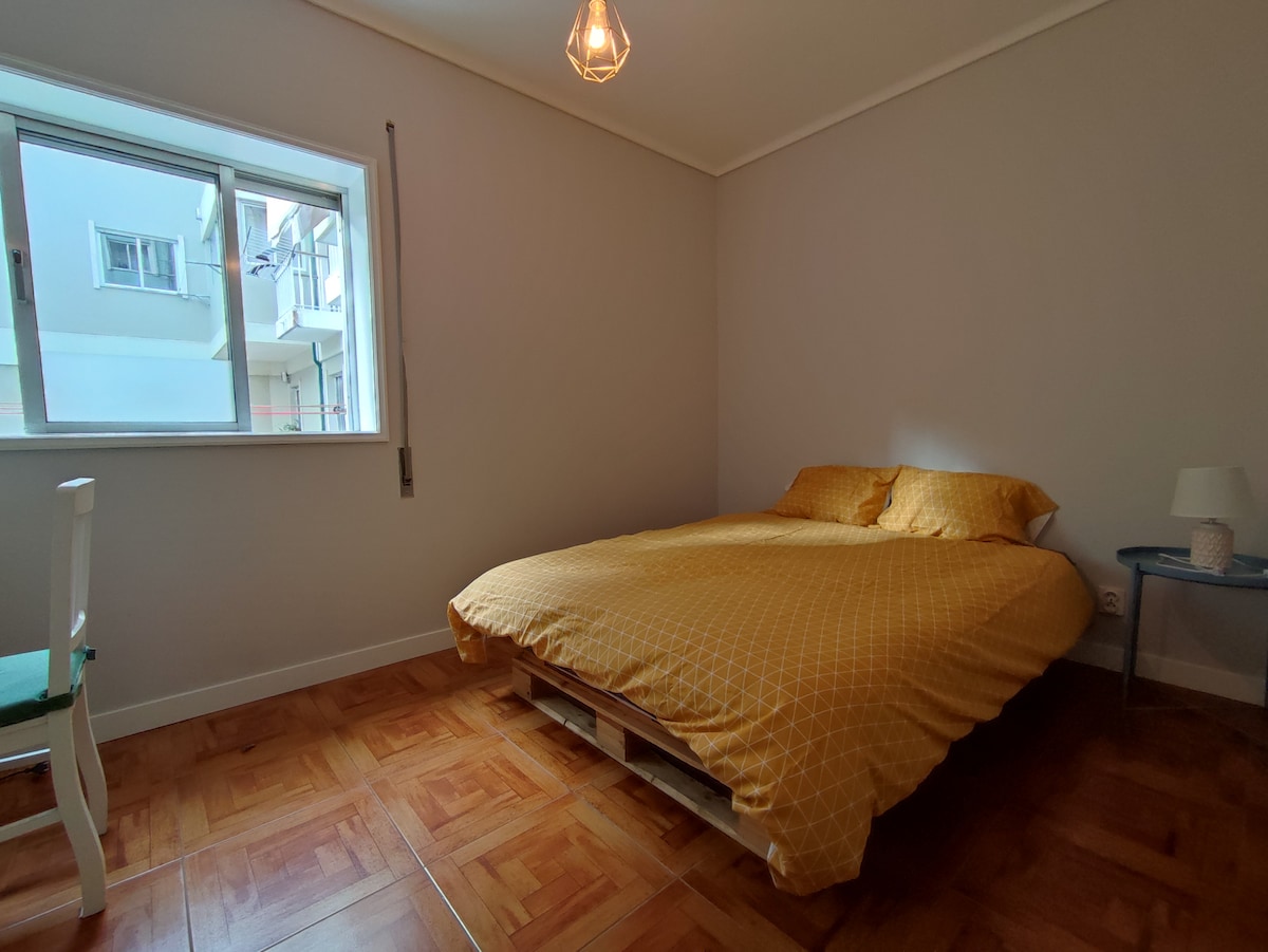 A bright and cozy room features a spacious bed covered with a yellow patterned comforter. Natural light fills the space through the large window, which overlooks the exterior. A simple side table and a chair add functionality to the minimalist design.
