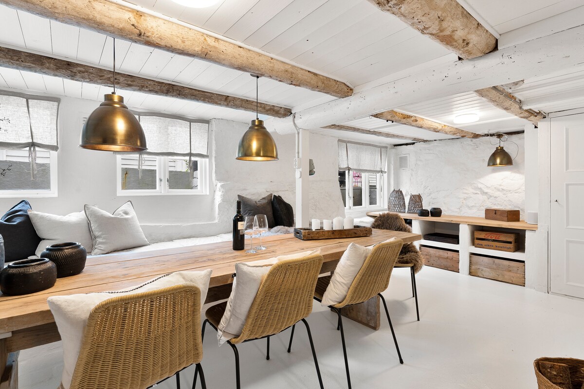 A spacious dining area is highlighted by a long wooden table surrounded by rattan chairs. Soft lighting is provided by pendant fixtures hanging from exposed beams. A cozy seating area features cushions on a built-in bench, with natural light filtering through the windows.