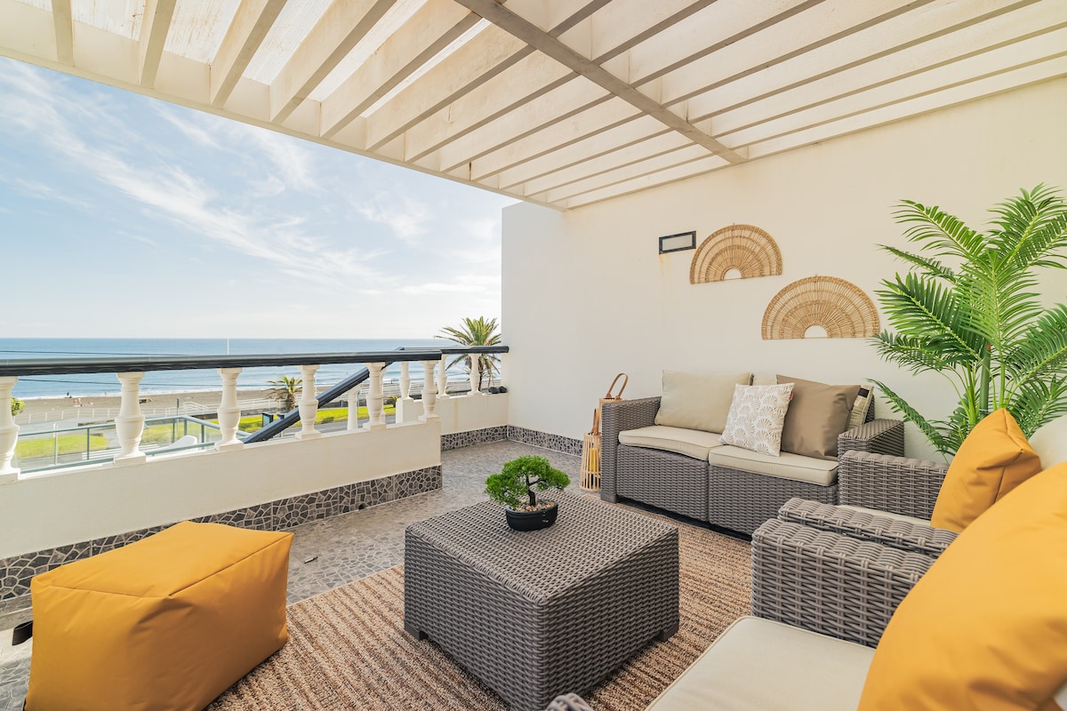 A spacious patio area features comfortable seating with light-colored cushions and decorative throw pillows. A coffee table made of woven material is centered on a natural fiber rug. Lush greenery and ocean views create a serene atmosphere, complemented by a pergola overhead allowing soft light to filter through.