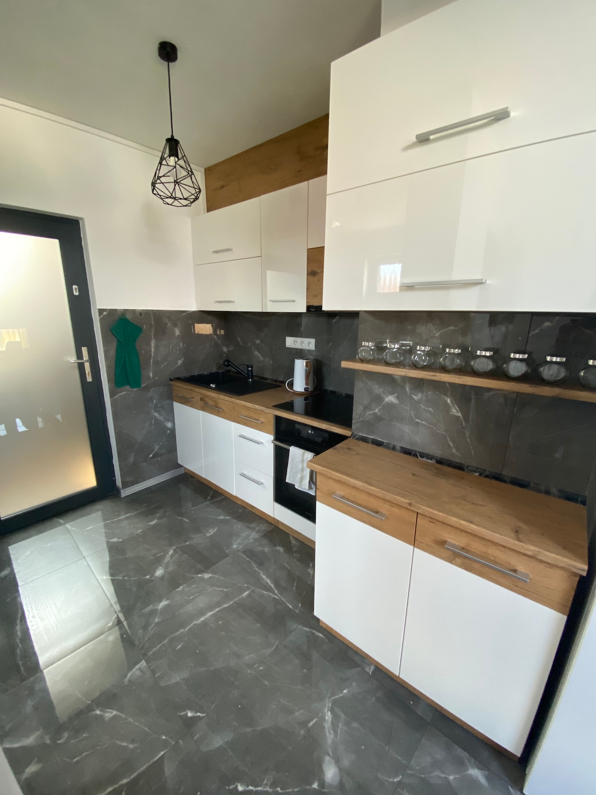 The kitchen showcases a modern design with a mix of white cabinetry and natural wood. Counter space is provided for meal preparation, while a glass set is neatly arranged above the counter, enhancing the functional layout. A pendant light adds subtle illumination.