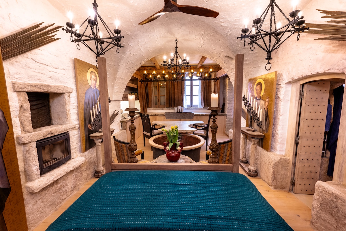 A cozy bedroom features a spacious canopied bed with a deep blue blanket. Surrounding the room, Byzantine icon art enhances the ambiance. A closed fireplace adds a touch of warmth, while arched ceilings and multiple light fixtures illuminate the space, creating a serene and inviting atmosphere.