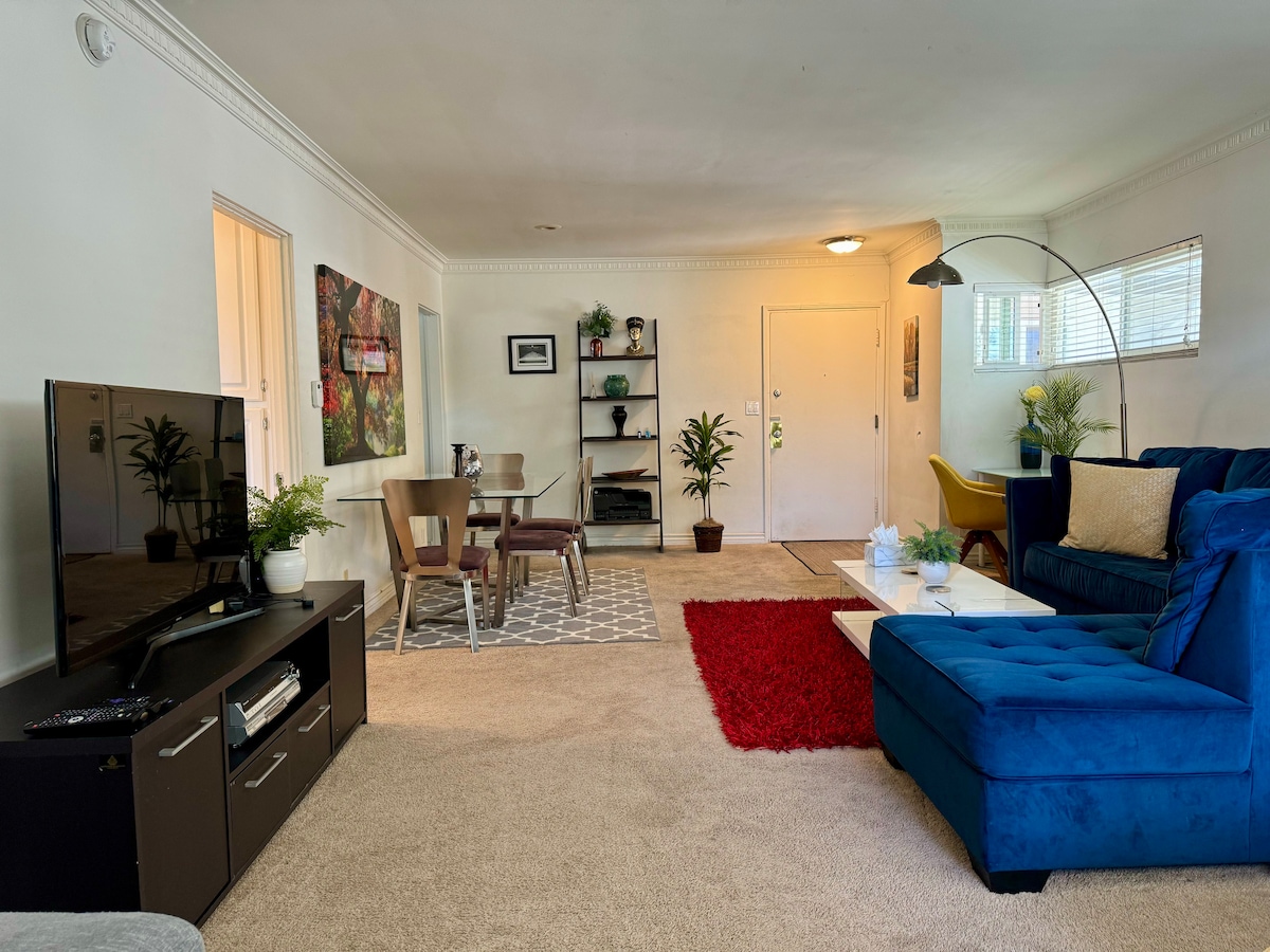 The living area features a deep blue sectional sofa and a flat-screen TV. A small dining set for four is situated nearby, along with a decorative rug. Natural light enters through abundant windows, and various plants decorate the space, enhancing the overall ambiance.