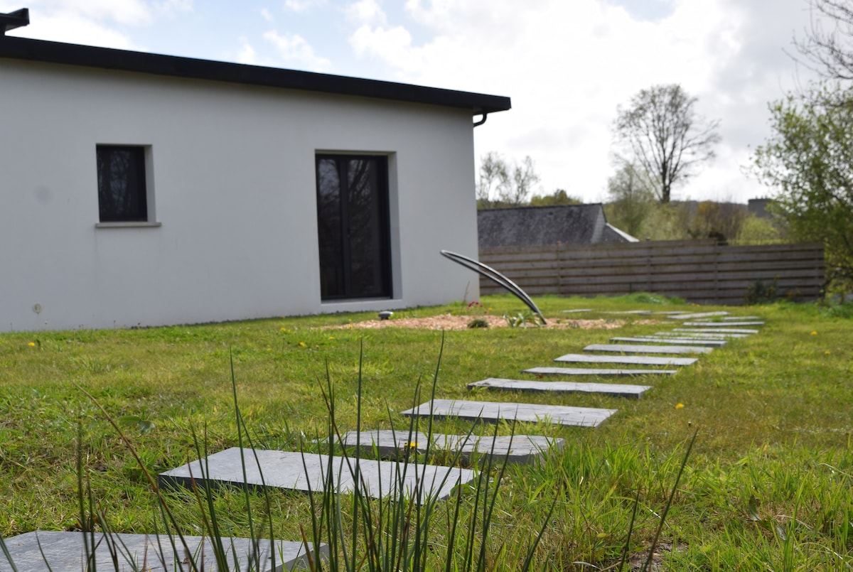 A private garden path leads to a modern white building with large windows. Stone slabs create a walkway through a grassy area, which is dotted with small plants. The surroundings feature trees and a wooden fence, offering a serene outdoor setting.