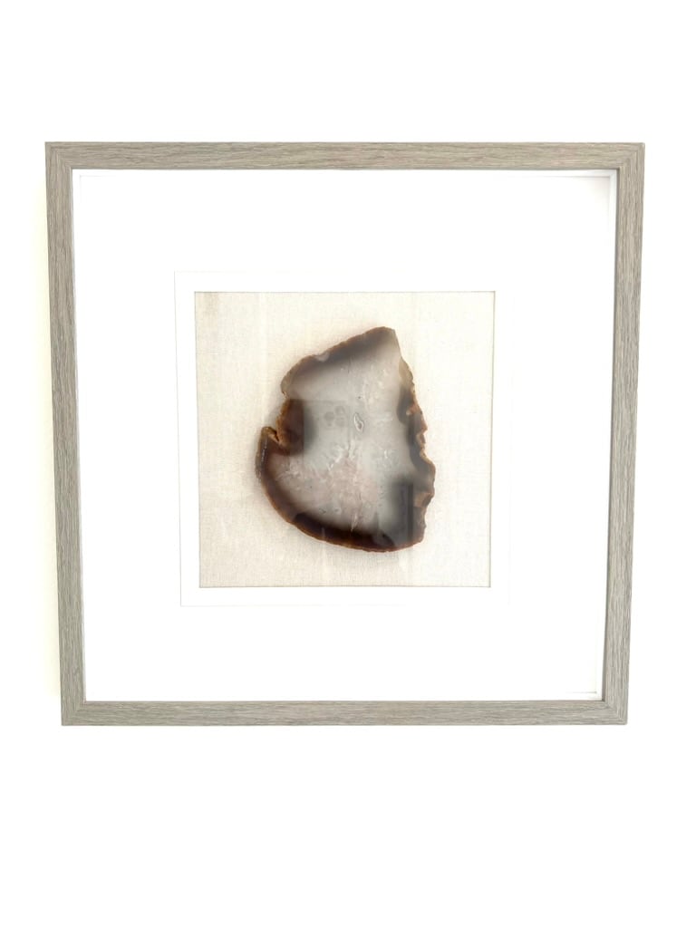 A framed wall art piece features a natural agate slice with rich browns and translucent sections. The artwork is set against a textured light background, enhancing its organic beauty. A simple, light wood frame complements the overall design.