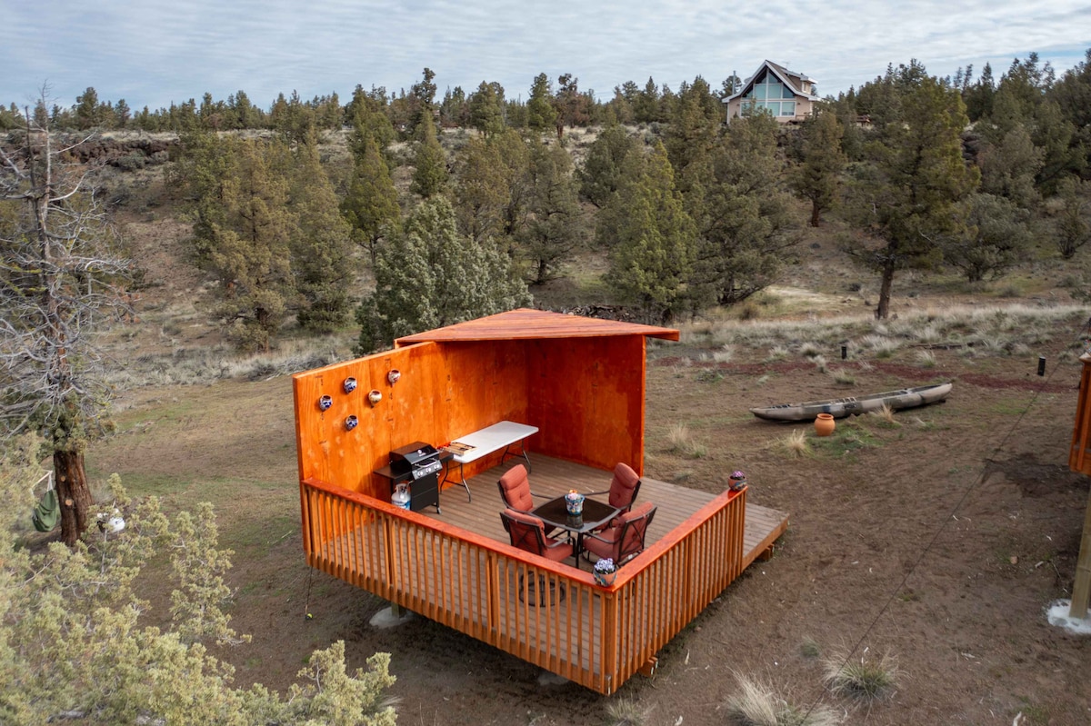 An outdoor lounge deck features orange-painted walls, furnished with two chairs and a table. The setting is surrounded by low greenery and scattered trees, providing a rustic atmosphere. A canoe and additional outdoor decor can be seen in the background.
